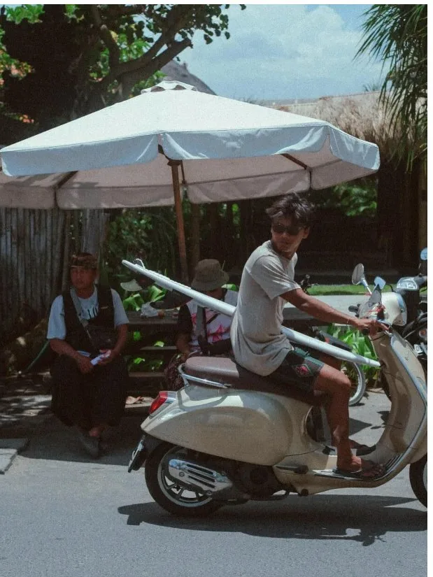 Surfer sitting on a scooter with surfboards nearby.